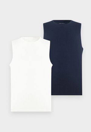 Homme portant un débardeur blanc, pantalon bleu marine, baskets blanches et casquette gris foncé, debout devant un fond blanc uni.