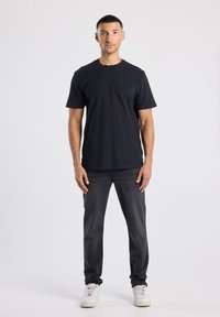 Camiseta de algodón negra con cuello redondo y mangas cortas, combinada con jeans de mezclilla gris oscuro y zapatillas blancas, de pie contra un fondo neutro.