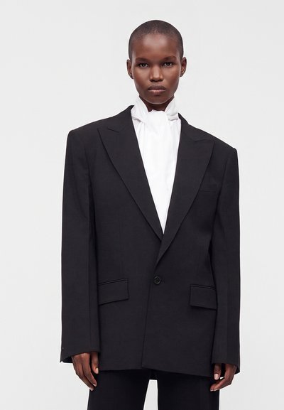 Personne aux cheveux courts portant un blazer noir oversize et un chemisier blanc à col montant sur un fond clair uni.