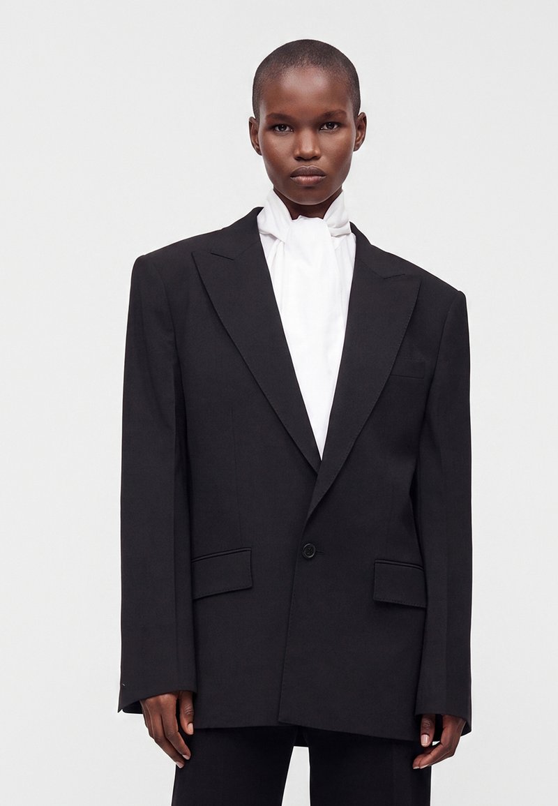 Personne aux cheveux courts portant un blazer noir oversize et un chemisier blanc à col montant sur un fond clair uni.