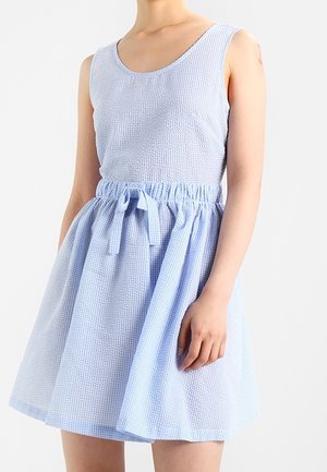 Bezrękawna, jasnoniebieska sukienka o fakturowanej powierzchni, z okrągłym dekoltem i marszczoną talią, zawiązana materiałową kokardą, noszona przez stojącą osobę na jednolitym tle.