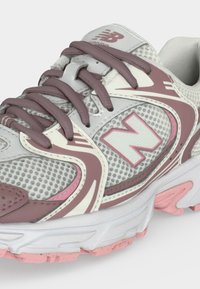 Κοντινό πλάνο ενός γκρι και ροζ παπουτσιού New Balance για τρέξιμο με διχτυωτό ύφασμα, λογότυπο στο πλάι και ροζ κορδόνια.
