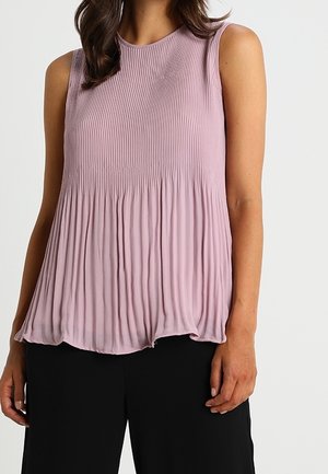 Mujer con blusa sin mangas plisada de color lila claro y pantalones negros sobre un fondo liso.