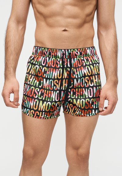 Torse masculin portant un short de bain coloré avec le lettrage répété "MOSCHINO" et un cordon noir sur un fond blanc.