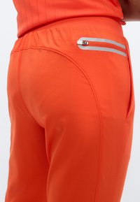 Orange atletikbukser lavet af glat stof, med en sidelomme med lynlås og sølvfarvede detaljer. Linningen er rynket for komfort.