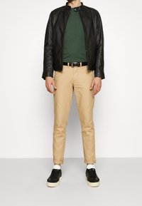 Černá kožená bunda, zelené tričko, béžové chinos kalhoty a černé sneakers s bílými podr�ážkami. Bunda má kulatý límec a přední zip.