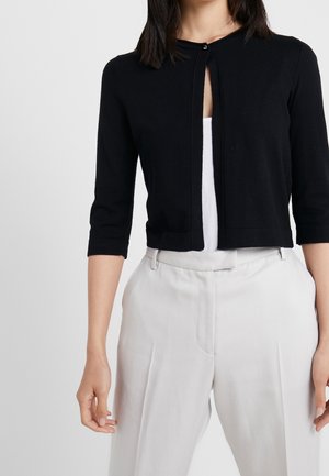 Femme portant un cardigan court noir avec un seul bouton supérieur, par-dessus un haut blanc et un pantalon blanc taille haute, debout devant un fond uni.