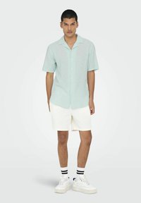 Camisa de manga corta azul clara con diseño de botones, combinada con pantalones cortos blancos texturizados y zapatillas blancas con detalles en negro.