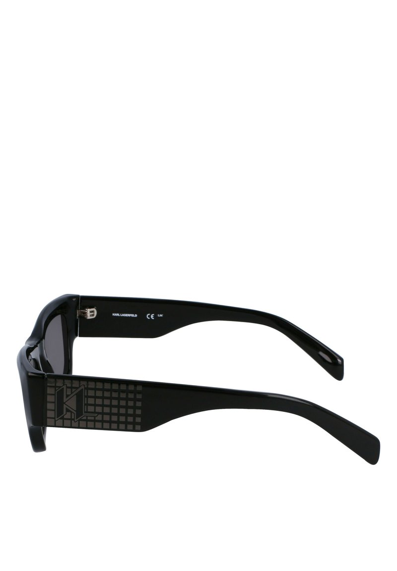 Lunettes noires avec des verres rectangulaires, monture épaisse, branches noires texturées avec un motif en grille, et un logo discret sur le côté.