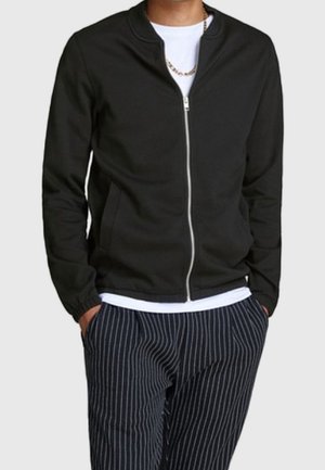 Mężczyzna w czarnej kurtce na zamek błyskawiczny noszonej na białej koszuli, z rękami w kieszeniach ciemnych spodni w prążki.