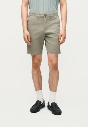 Man draagt beige korte broek tot aan de knie, witte crew sokken, zwarte instapschoenen en een mouwloos lichtgrijs geribbeld topje.