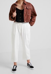 Giacca di jeans corta marrone con bottoni, top nero con bottoni, pantaloni bianchi a vita alta e sneakers nere con dettagli bianchi.