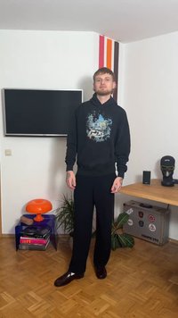 Zwarte hoodie met een grafisch ontwerp van landschappen, gecombineerd met donkere broek en glanzende bruine schoenen. Binnenomgeving met kleurrijke decoratie.