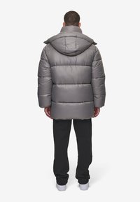 Grå pufferjakke med hette, med en quiltet design og en avslappet passform. Bæret over svarte bukser og hvite joggesko.