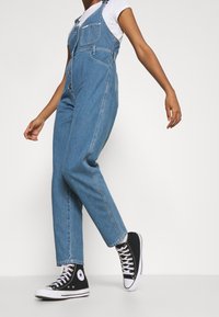 Ljusblå denimoveraller med avslappnad passform, som har justerbara axelremmar, framfickor och är kombinerade med svarta höga sneakers.