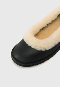 Pantofole in pelle nera con un rivestimento in pile crema attorno all'apertura. Sole piatto con sottostrato testurizzato per una buona aderenza. Design semplice e minimalista.