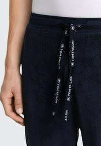 Gros plan sur une personne portant un pantalon de survêtement bleu marine avec un cordon de serrage imprimé "Tom Tailor" et une chemise blanche.