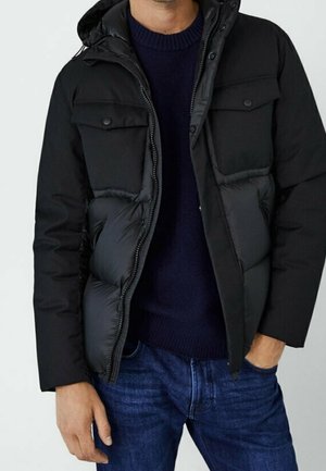 Homme portant une veste matelassée noire avec poches à rabat et capuche, par-dessus un pull bleu marine et un jean bleu.