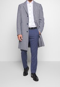 Houndstooth patterned coat in black and white, buttoned with blue accents. Underneath, a white shirt and blue checkered trousers, paired with black shoes.