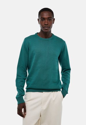 Pull turquoise à col rond en tissu tricoté, avec des manches longues et des poignets côtelés, associé à un pantalon clair à coupe ample.