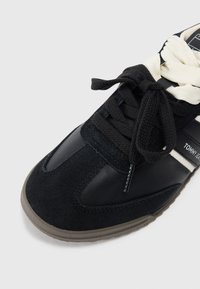 Svart sneakers med en kombinasjon av semsket skinn og lær, kremfargede lisser og en gummisåle med et teksturert mønster.