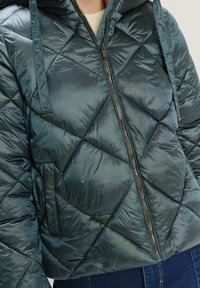 Pružná bunda v tmavom teal s prešívaným diamantovým vzorom, so zipsom a kapucňou; vyrobená z lesklého, ľahkého materiálu.