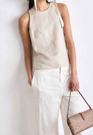 Kobieta w beżowym topie bez rękawów i białych spodniach z szerokimi nogawkami, trzymająca brązową torebkę w wzory z cienkim paskiem na ramię.
