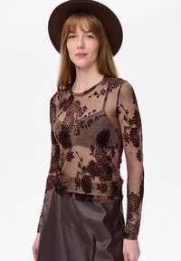 Vrouw met een brede bruine hoed, doorschijnende lange mouwen met bloemenpatroon, een bruine bralette en een bruine leren rok, poseert tegen een witte achtergrond.