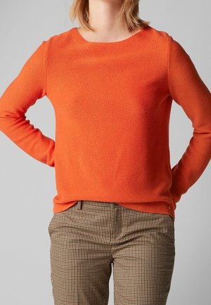 Osoba ubrana w jasnopomarańczowy sweter z długim rękawem i brązowe spodnie w kratę, z rękami opartymi na biodrach na jednolitym tle.