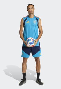 Blue athletic jersey with sleeveless design, paired with navy shorts featuring red and blue accents. Holding a multicolored soccer ball.