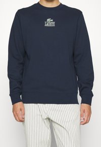 Sudadera de algodón azul marino con mangas largas, puños acanalados y cuello redondo; presenta un logo bordado de Lacoste en el pecho.