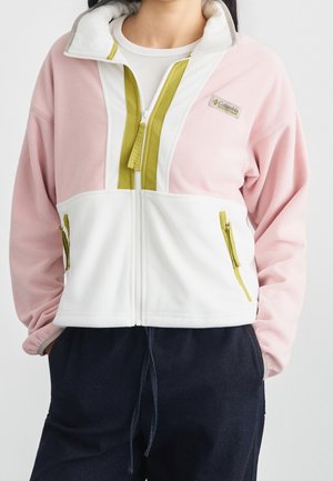 Vrouw draagt een pastelroze en witte fleecejas met een rits met olijfgroene ritssluitingen en donkerblauwe broek, handen in de zakken.
