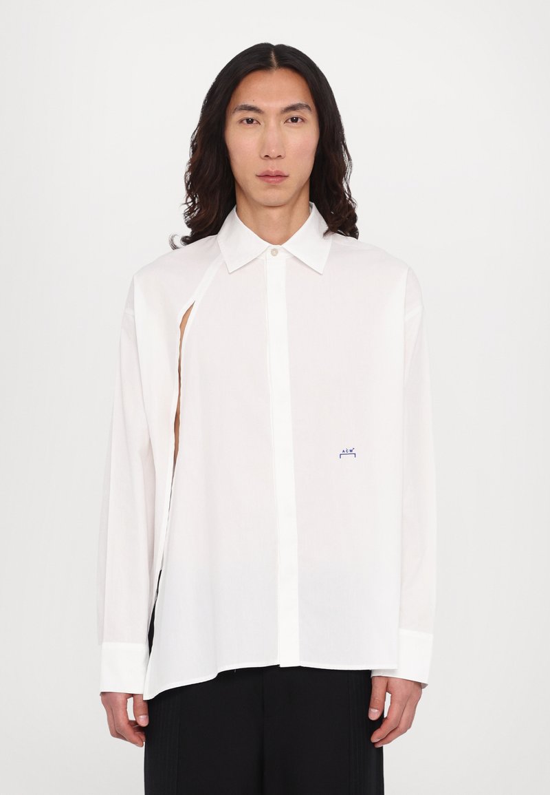 Chemise blanche à manches longues avec col pointu, présentant une découpe sur le côté et un petit logo bleu. Tissu frais avec fermeture à boutons.