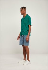 Groene korte mouw knoopschema met één borstzak, gecombineerd met lichtblauwe denim shorts en witte sneakers. Eenvoudig ontwerp en ontspannen pasvorm.