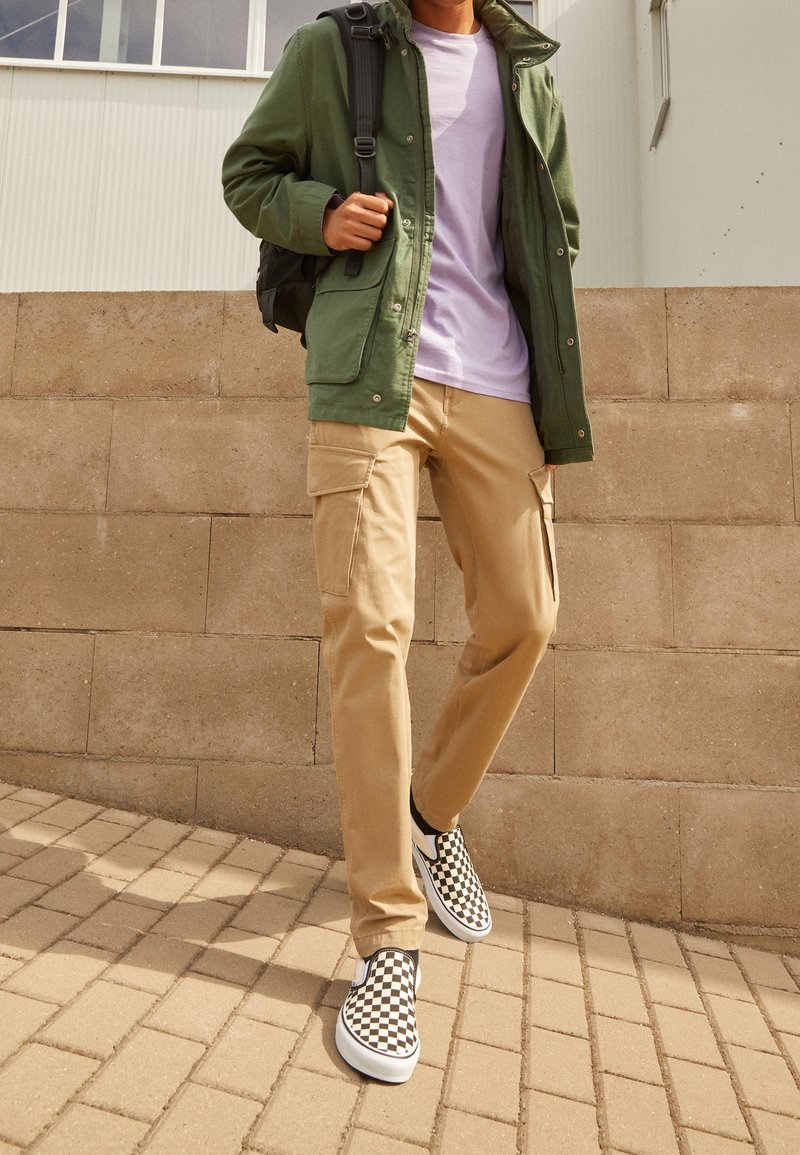 Personne marchant à l'extérieur portant un pantalon cargo beige, une veste verte, une chemise violette, des chaussures slip-on à damiers et un sac à dos noir.