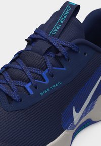 Εγγύς λήψη ναυτικού μπλε παπουτσιού τρεξίματος Nike Trail με κορδόνια μπλε με στίγματα, "Juniper Trail" στη γλώσσα και λευκό λογότυπο Nike στο πλέγμα της πλευράς.