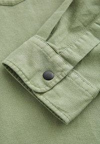 Tessuto verde chiaro con texture e risvolto. Presenta un dettaglio in bottone nero. Il materiale appare morbido e intrecciato.