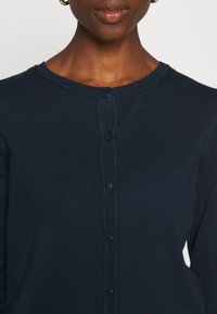 Marineblauwe gebreide cardigan met ronde halslijn, voorzien van zes donkere knopen aan de voorkant en ribbelstructuur langs de randen.