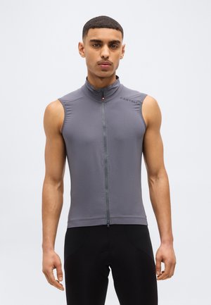 Férfi szürke, ujjatlan, cipzáras kerékpáros mellényt és fekete, testhez simuló nadrágot viselve, sima világos háttér előtt áll.