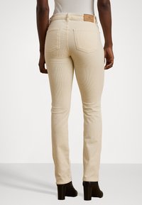 Beige corduroy jeans feature a high waist, back pockets, and a branded leather patch on the waistband. The fit is slim and streamlined.