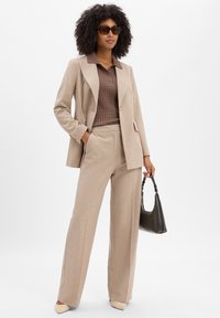 Vrouw in een beige blazer en wijde broek, bruin gebreid polo, beige hakken, zonnebril en een zwarte handtas met patroon.