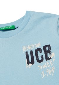 Jasnoniebieska koszulka z krótkim rękawem i okrągłym dekoltem. Posiada grafikę w ciemnoniebieskim i białym kolorze, w tym napis "BENETTON" oraz "UCB since 1965."