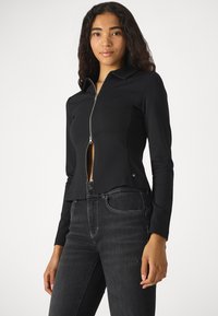 Veste noire zippée avec des manches longues et un col haut. Tissu lisse avec une coupe ajustée, associée à un jean foncé taille haute avec des coutures visibles.