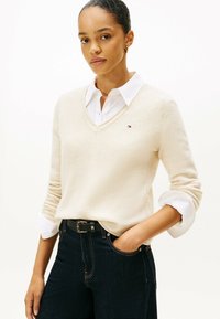 Vrouw met een opgetrokken haar in een crèmekleurige V-hals trui over een wit shirt, donkere jeans en een zwarte riem, poserende met één hand in de broekzak.