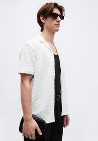 Άνδρας με μαύρα γυαλιά ηλίου φοράει υφασμένο λευκό κοντομάνικο πουκάμισο πάνω από μαύρο τοπ και κρατάει μικρό μαύρο υφασμένο τσαντάκι χειρός.