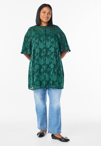 Tmavozelená blúzka s kvetinovým vzorom a krátkymi rukávmi, kombinovaná so svetlomodrými džínsami s širokými nohavicami a čiernymi plochými topánkami. Jemná, štruktúrovaná látka.