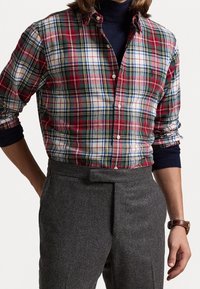 Camicia a quadri rossa e verde con accenti bianchi e blu, chiusa con bottoni. Maniche lunghe arrotolate, abbinata a pantaloni grigi su misura.