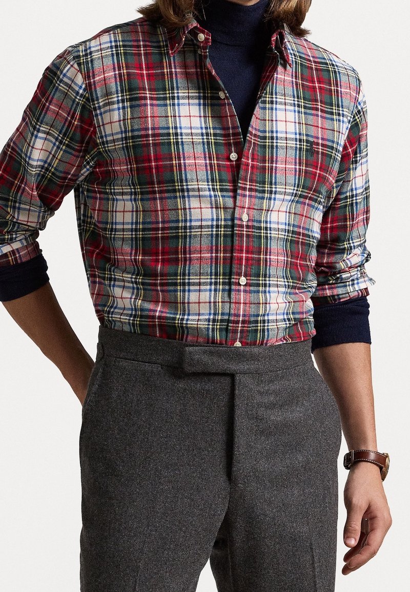 Red and green plaid button-up shirt with white and blue accents. Long sleeves rolled up, paired with gray tailored trousers.