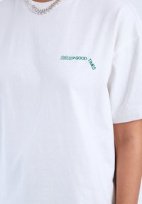Bílý bavlněný tričko s zeleným potiskem textu "Dr. Drit" a "GOOD TIMES" v úhlu. Krátké rukávy a volný střih.