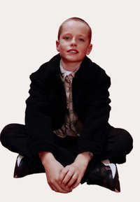 Sort, fleece-fórret jakke over en ternet skjorte, sorte bukser og sorte sneakers. Siddeposition med hænderne foldet på knæene. Neutral baggrund.